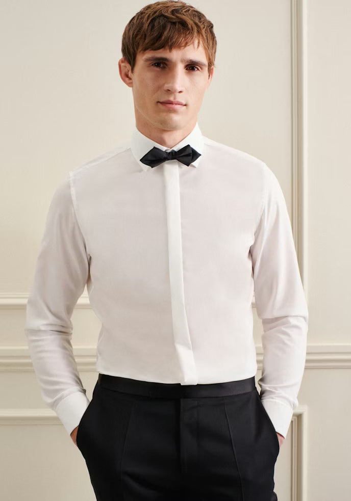 Man wearing a white dress shirt with a black bow tie and black pants against a neutral wall.