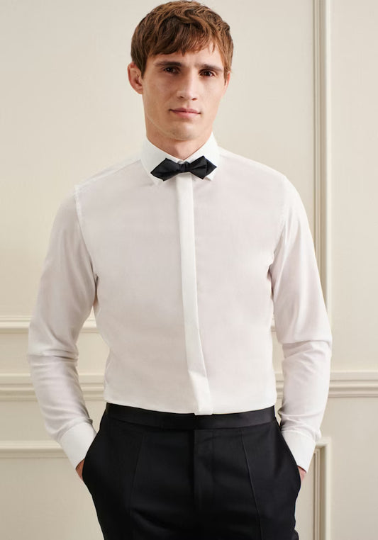 Man wearing a white dress shirt with a black bow tie and black pants against a neutral wall.
