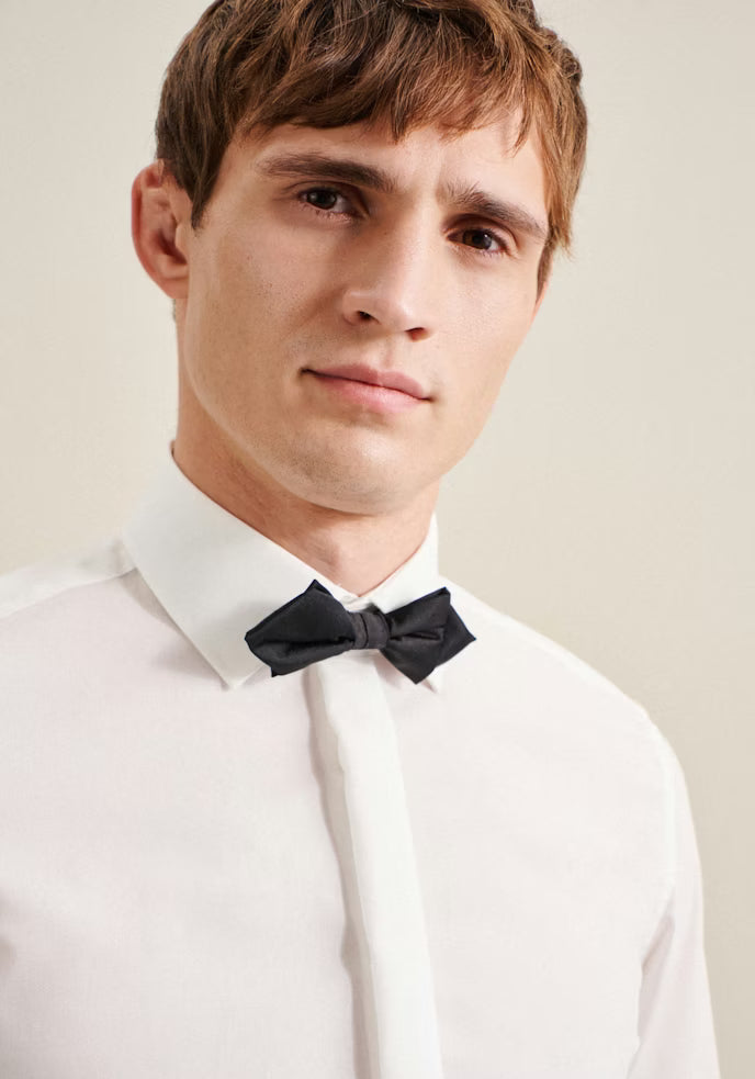 Man wearing a white shirt with a black bow tie against a plain background