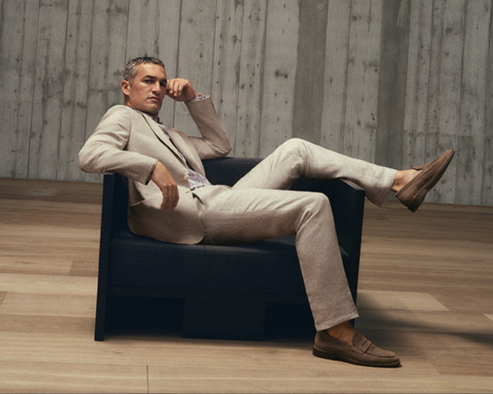 Man in a beige suit sitting on a black chair against a concrete wall.