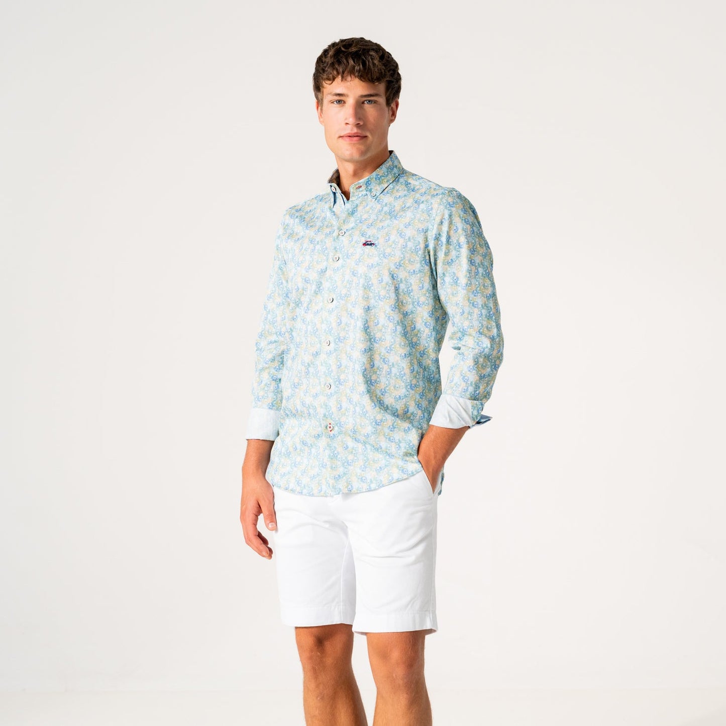 A man modeling a light blue and white patterned long-sleeve button-down shirt with white shorts, standing against a plain white studio background.