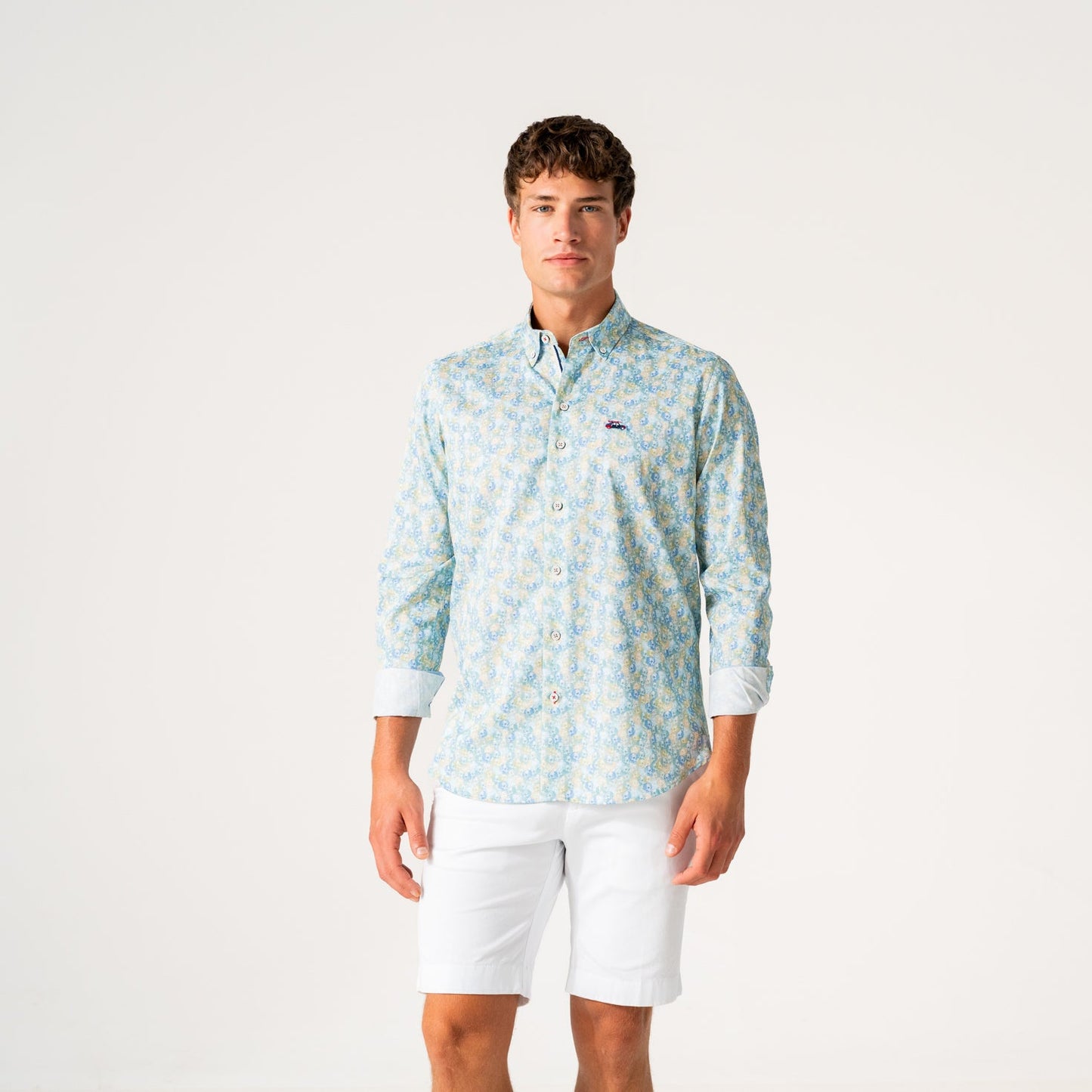 A young man modeling a light blue and white patterned long-sleeve button-down shirt with white shorts, standing against a plain white studio background.