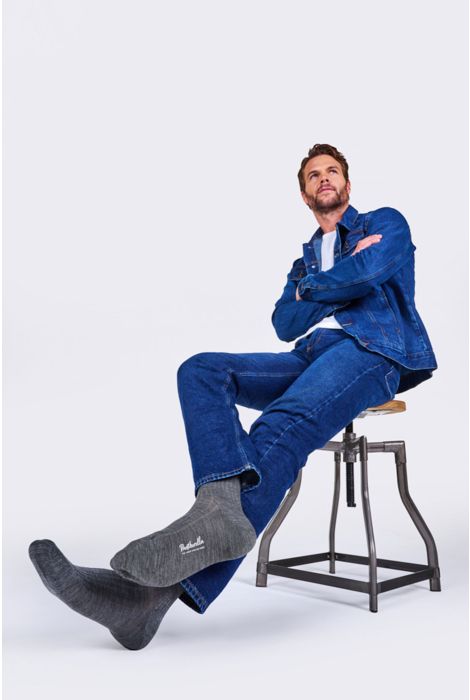 Man sitting on a stool wearing a denim outfit with gray socks against a white background