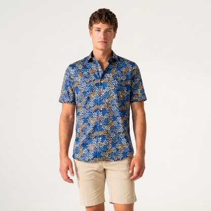 A man modeling a short-sleeve button-up shirt with a blue, gold, and white floral pattern, paired with beige shorts, against a plain white studio background.
