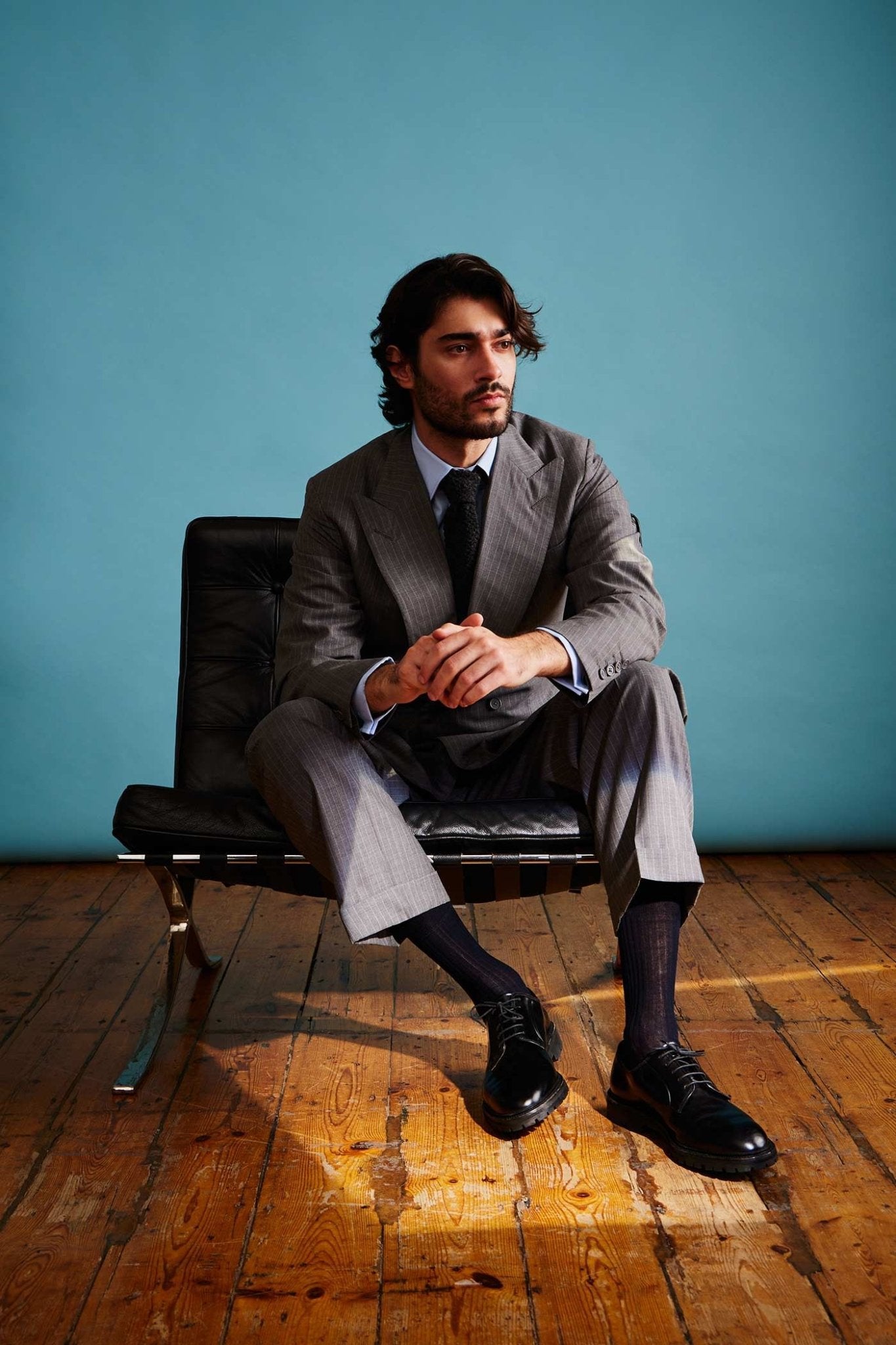 Man in a gray suit sitting on a black chair against a blue wall.