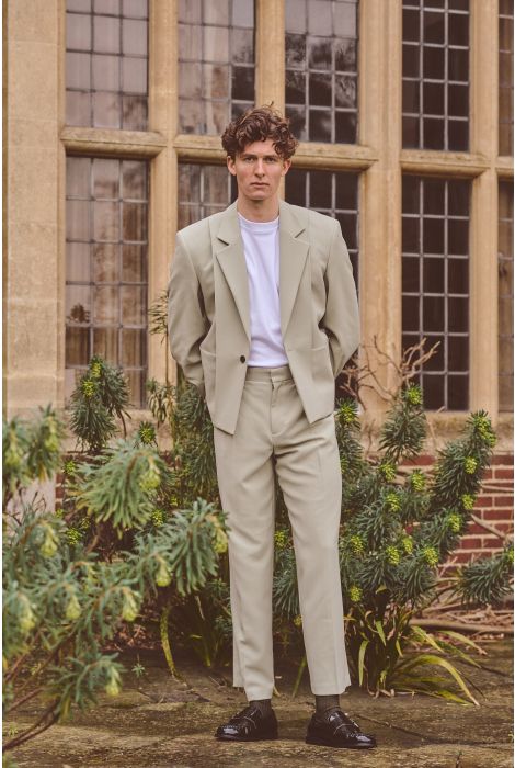 Man in a beige suit standing in front of a building with large windows and greenery.