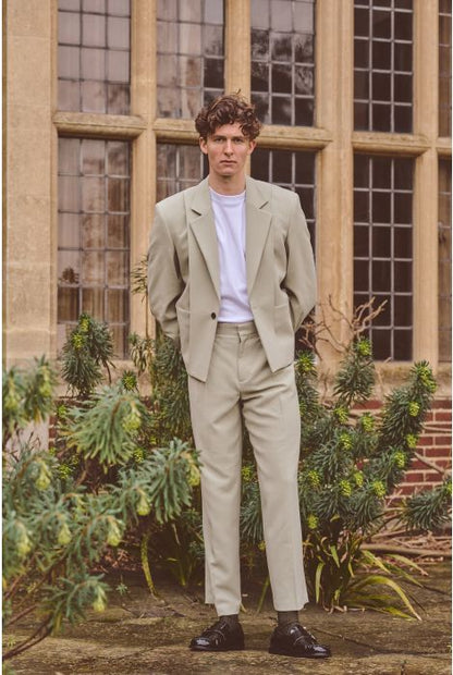 Man in a beige suit standing in front of a building with large windows and greenery.